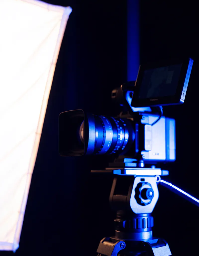 Escena ilustrativa: una camara de cine montada en tripie junto a un panel difusor iluminado, sobre fondo oscuro.