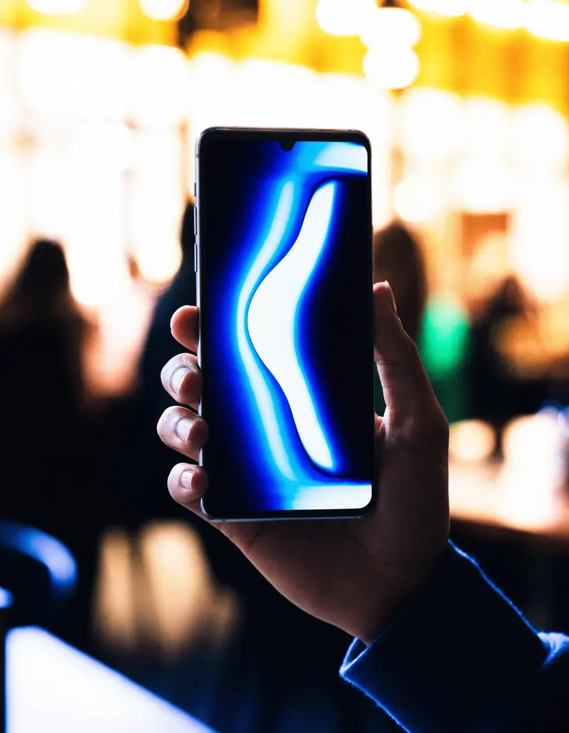 Escena ilustrativa: una mano sostiene un telefono en vertical reproduciendo una pieza abstracta, con luces desenfocadas al fondo.
